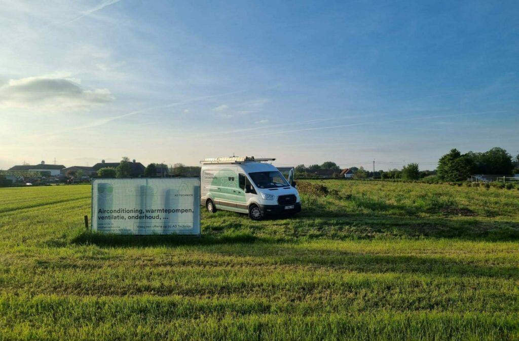 AD Technics - Een wit busje met het opschrift 'Over AD Technics' staat geparkeerd op een grasveld naast een bord met tekst over airconditioning, warmtepompen, ventilatie en onderhoud. Nabijgelegen gebouwen en bomen zijn zichtbaar onder een helderblauwe lucht.