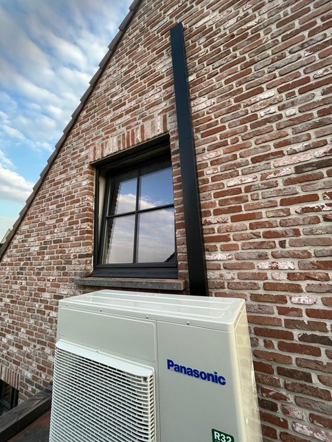 AD Technics - Een Panasonic airconditioning unit is geïnstalleerd onder een raam op een bakstenen muur van een huis. Een verticale zwarte leiding is bevestigd naast het raam, waardoor het een uitstekende keuze is voor degenen die overwegen om warmtepompen te kiezen.